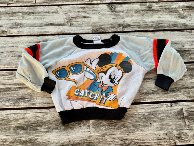 May include: A white long-sleeve sweatshirt featuring a graphic of Mickey Mouse holding yellow sunglasses with the text CATCH IT. The garment has light gray sleeves with black and red stripes at the shoulders, black cuffs, and a black crew neckline.