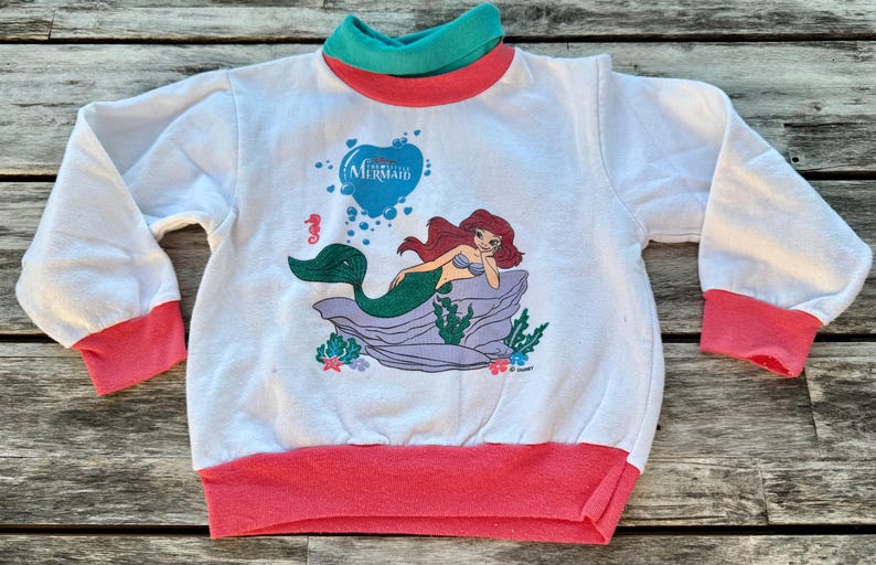 Op de afbeelding: Een wit sweatshirt met lange mouwen en een afbeelding van de kleine zeemeermin Ariel op een rots. Het kledingstuk heeft een blauwgroene opstaande kraag, koraalroze mouwboorden en een koraalroze zoom. De tekst The Little Mermaid staat erboven.