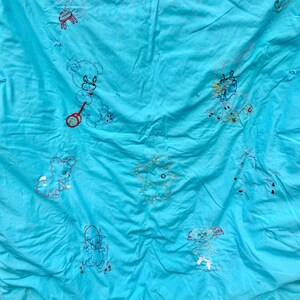 May include: A vintage turquoise quilt with embroidered teddy bear designs in various poses. The bears are outlined in red, black, and white thread, with some holding toys. The quilt has a crinkled texture and appears to be handmade.