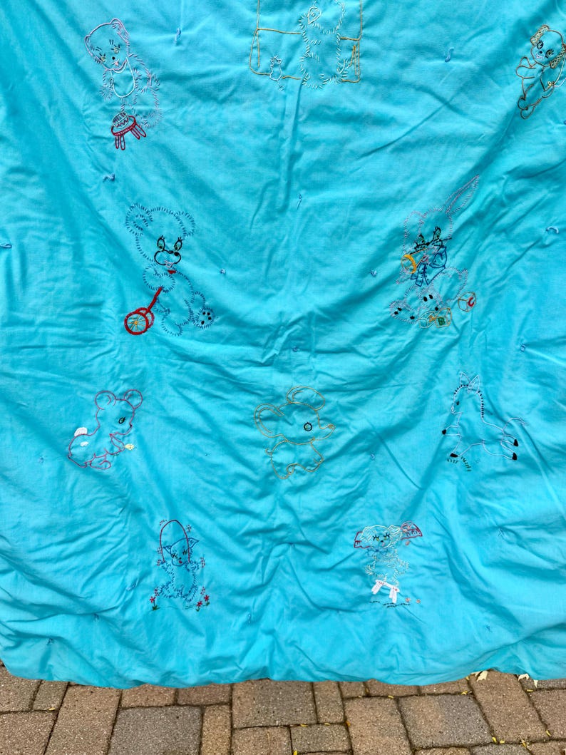 May include: A vintage turquoise quilt with embroidered teddy bear designs in various poses. The bears are outlined in red, black, and white thread, with some holding toys. The quilt has a crinkled texture and appears to be handmade.