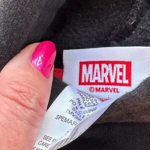 May include: A black garment featuring a white fabric tag with the red Marvel logo and the text MARVEL printed below it. A secondary white care label is attached, displaying text including PO, RN, and style number 7MV391, with Made in Tamil Nadu.