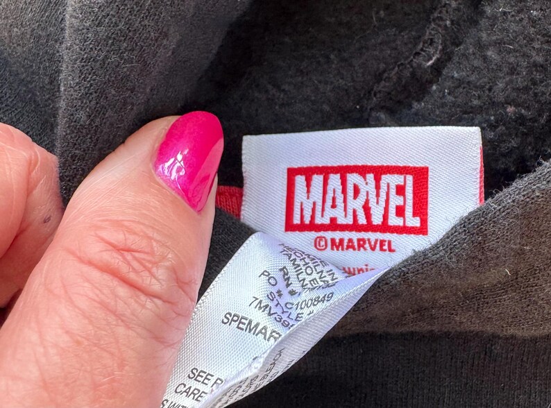 May include: A black garment featuring a white fabric tag with the red Marvel logo and the text MARVEL printed below it. A secondary white care label is attached, displaying text including PO, RN, and style number 7MV391, with Made in Tamil Nadu.