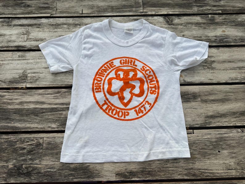May include: A white short-sleeve t-shirt featuring an orange circular graphic on the chest. The graphic displays the text "BROWNIE GIRL SCOUTS TROOP 1473" surrounding a central clover-shaped emblem with a stylized figure in the middle.