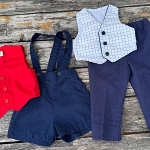 Puede incluir: Un chaleco rojo, unos pantalones cortos azul marino con tirantes, un chaleco blanco a cuadros y unos pantalones azul marino están dispuestos sobre una superficie de madera. El chaleco tiene tres botones. Los pantalones cortos tienen tirantes con botones.