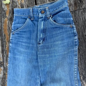 Puede incluir: Primer plano de unos vaqueros de mezclilla azul vintage. Los vaqueros tienen cierre de bot&oacute;n, bolsillos delanteros y un dise&ntilde;o cl&aacute;sico de pernera recta. La mezclilla tiene un aspecto descolorido, lo que sugiere desgaste y antig&uuml;edad.