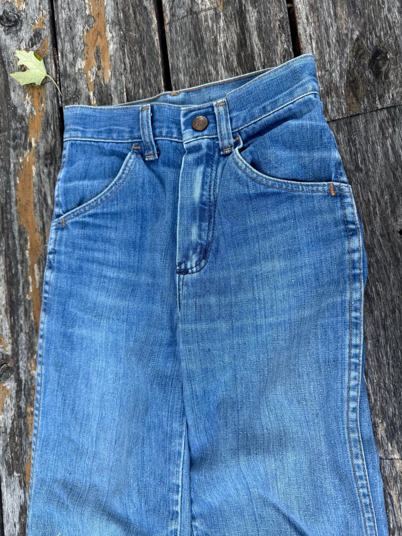 Puede incluir: Primer plano de unos vaqueros de mezclilla azul vintage. Los vaqueros tienen cierre de bot&oacute;n, bolsillos delanteros y un dise&ntilde;o cl&aacute;sico de pernera recta. La mezclilla tiene un aspecto descolorido, lo que sugiere desgaste y antig&uuml;edad.