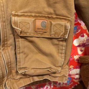 Puede incluir: Primer plano de un bolsillo de chaqueta Carhartt. El bolsillo es de color marr&oacute;n claro y tiene un parche cuadrado con el logotipo de Carhartt. La chaqueta est&aacute; hecha de una tela duradera y texturizada. El bolsillo es rectangular y tiene una solapa.