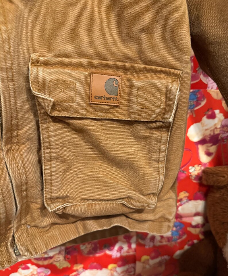 Puede incluir: Primer plano de un bolsillo de chaqueta Carhartt. El bolsillo es de color marr&oacute;n claro y tiene un parche cuadrado con el logotipo de Carhartt. La chaqueta est&aacute; hecha de una tela duradera y texturizada. El bolsillo es rectangular y tiene una solapa.