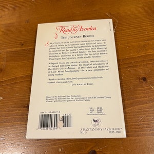 May include: A paperback book cover for "Road to Avonlea: The Journey Begins" based on the television series. The cover features a red title with a white background and a black and white illustration of a girl in a dress and a boy in a suit standing on a path.