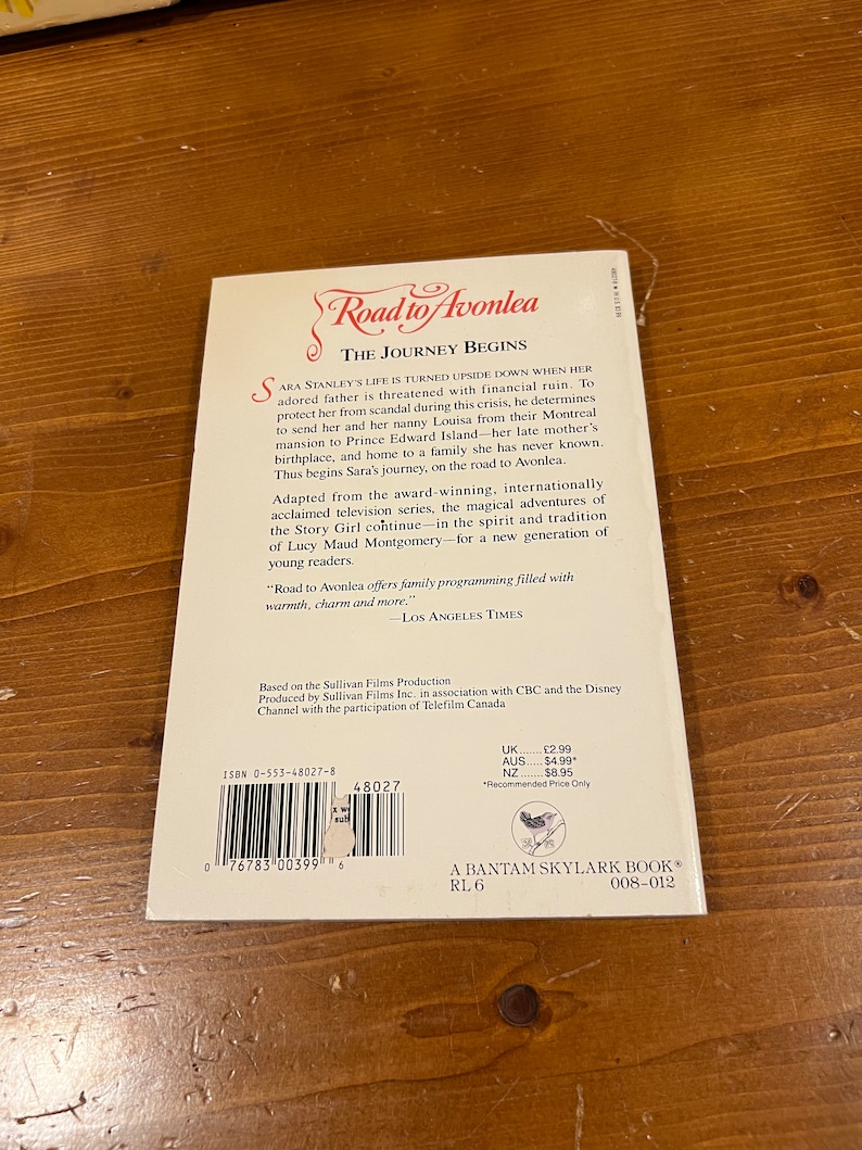 May include: A paperback book cover for "Road to Avonlea: The Journey Begins" based on the television series. The cover features a red title with a white background and a black and white illustration of a girl in a dress and a boy in a suit standing on a path.