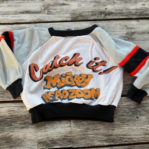 May include: A white long-sleeve sweatshirt with light gray sleeves, black cuffs, and a black collar. The front features the orange and blue text "Catch it! Mickey HEADZOOM" with red and black stripes on the sleeves near the shoulders.