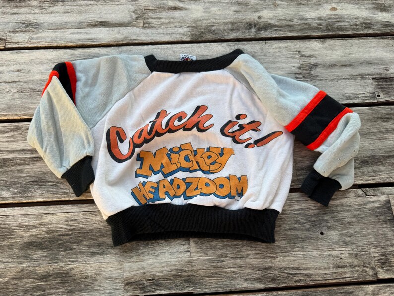 May include: A white long-sleeve sweatshirt with light gray sleeves, black cuffs, and a black collar. The front features the orange and blue text "Catch it! Mickey HEADZOOM" with red and black stripes on the sleeves near the shoulders.