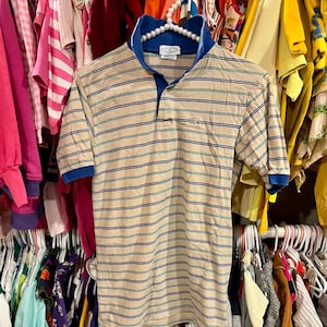 Pode incluir: Uma camisa polo bege listrada com listras azuis, verdes e brancas. A camisa tem um colarinho azul e fecho de botões. O logotipo do crocodilo Lacoste é bordado no peito esquerdo.