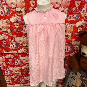 May include: A light pink sleeveless nightgown with a ruffled collar and lace trim. The nightgown features a small embroidered floral design and two buttons on the bodice. The fabric has a textured appearance.