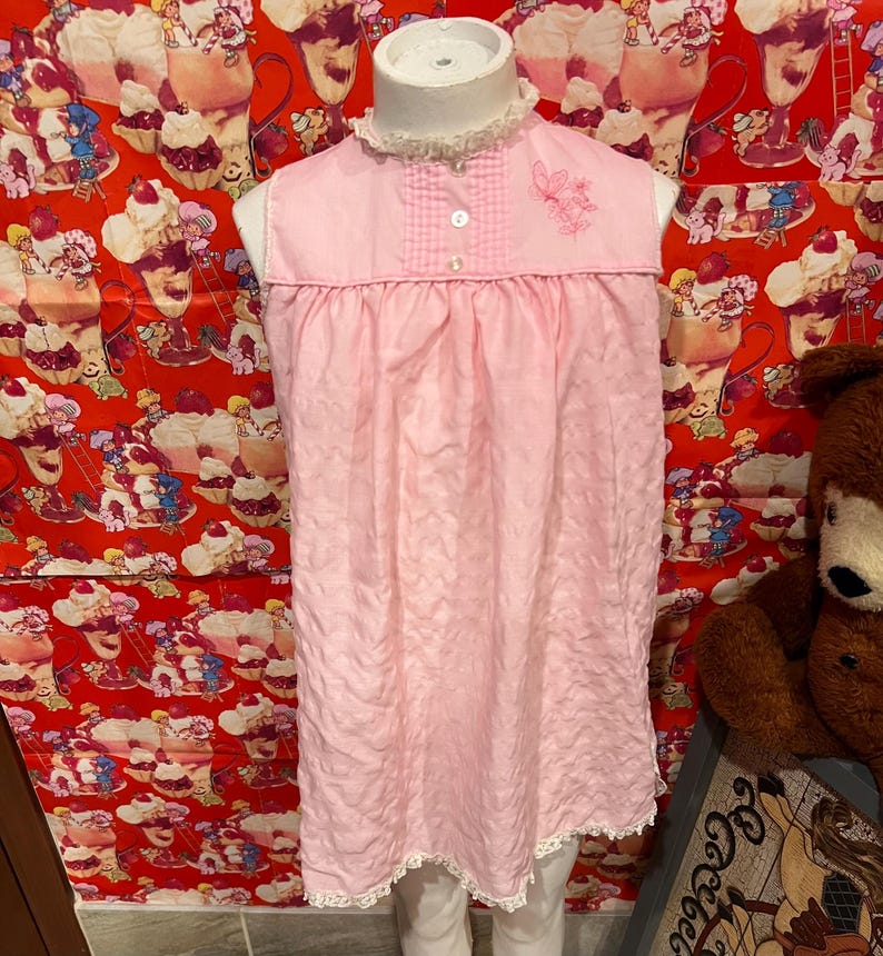 May include: A light pink sleeveless nightgown with a ruffled collar and lace trim. The nightgown features a small embroidered floral design and two buttons on the bodice. The fabric has a textured appearance.