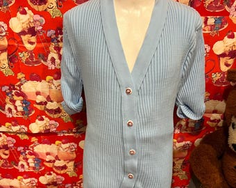 Könnte beinhalten: Hellblau gestrickte Rippstrickjacke mit V-Ausschnitt und kurzen Ärmeln. Die Strickjacke hat fünf dekorative Knöpfe mit floralem Design. Die Ärmel sind hochgekrempelt. Der Hintergrund ist ein rot-weiß gemusterter Stoff.