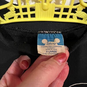 Op de afbeelding: Zwart sweatshirt met een witte Mickey Mouse-afbeelding en een blauw Disney Character Fashions-label. Het label vermeldt "Disney Character Fashions", "Disney", "Made in U.S.A.", "X-Large" en "Care & Fiber See".