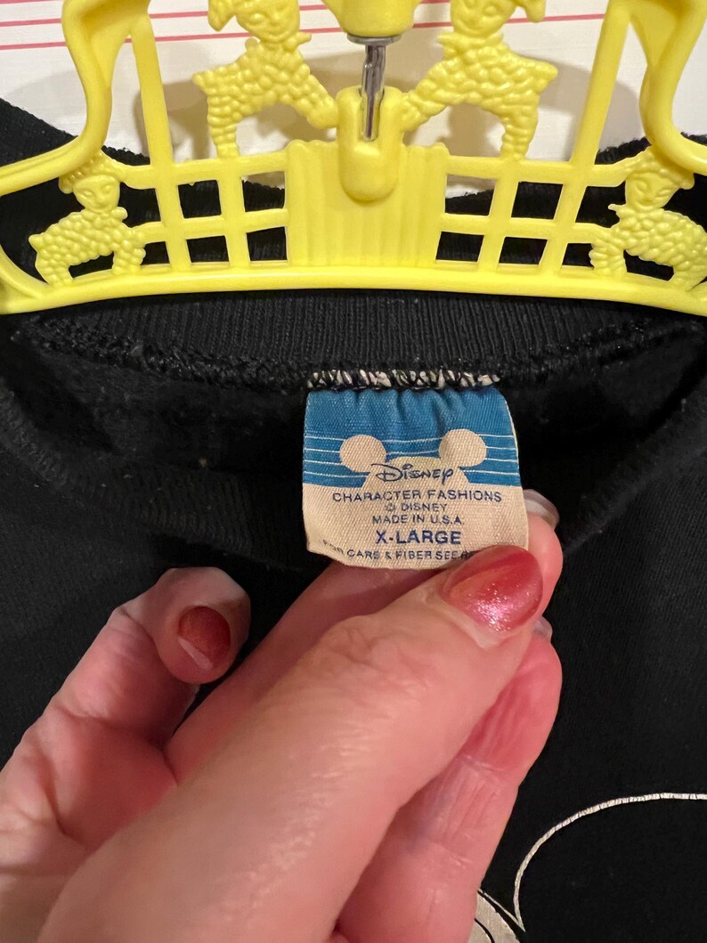 Op de afbeelding: Zwart sweatshirt met een witte Mickey Mouse-afbeelding en een blauw Disney Character Fashions-label. Het label vermeldt "Disney Character Fashions", "Disney", "Made in U.S.A.", "X-Large" en "Care & Fiber See".