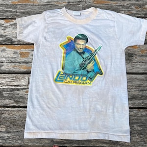May include: White t-shirt with a graphic of Lando Calrissian from Star Wars. The graphic is in a yellow and blue frame with the character's name below. The shirt has a light beige collar and shows signs of wear.