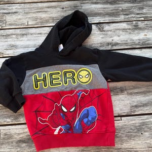May include: A long-sleeve hooded sweatshirt featuring a color-blocked design in black, gray, and red. The gray chest panel displays the yellow text HERO with a spider icon, while the red lower section features a graphic of the character Spider-Man.