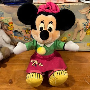 May include: A plush Minnie Mouse toy wearing a green dress, pink skirt, and yellow shoes. She has a pink bow in her hair and a yellow pom-pom on her dress.