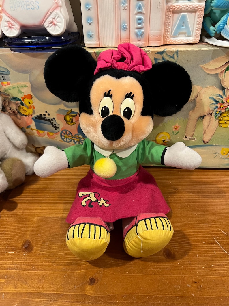 May include: A plush Minnie Mouse toy wearing a green dress, pink skirt, and yellow shoes. She has a pink bow in her hair and a yellow pom-pom on her dress.