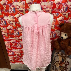 May include: A light pink vintage nightgown with a high neckline and a ruffled collar. The sleeveless dress has a gathered waist and a lace trim along the hemline. The back features a button closure. The nightgown is displayed on a mannequin.