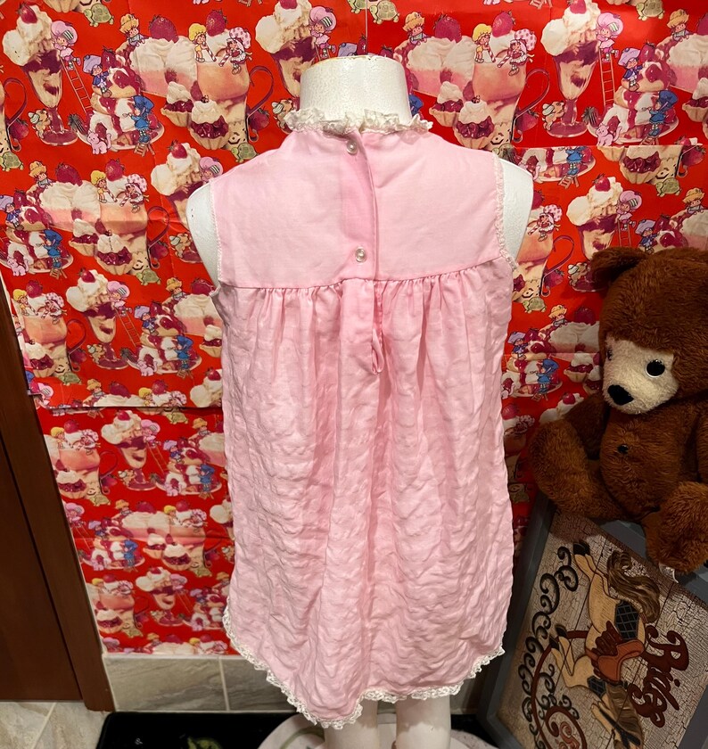 May include: A light pink vintage nightgown with a high neckline and a ruffled collar. The sleeveless dress has a gathered waist and a lace trim along the hemline. The back features a button closure. The nightgown is displayed on a mannequin.