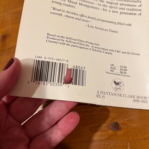 May include: A white book with a black and white barcode, the title "Road to Avonlea" and the ISBN 0-553-48027-8. The book is a Bantam Skylark book with a reading level of 6. The back cover features a small bird illustration.