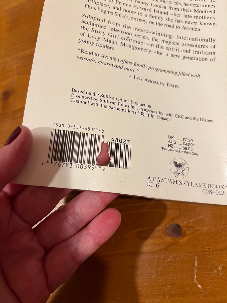 May include: A white book with a black and white barcode, the title "Road to Avonlea" and the ISBN 0-553-48027-8. The book is a Bantam Skylark book with a reading level of 6. The back cover features a small bird illustration.