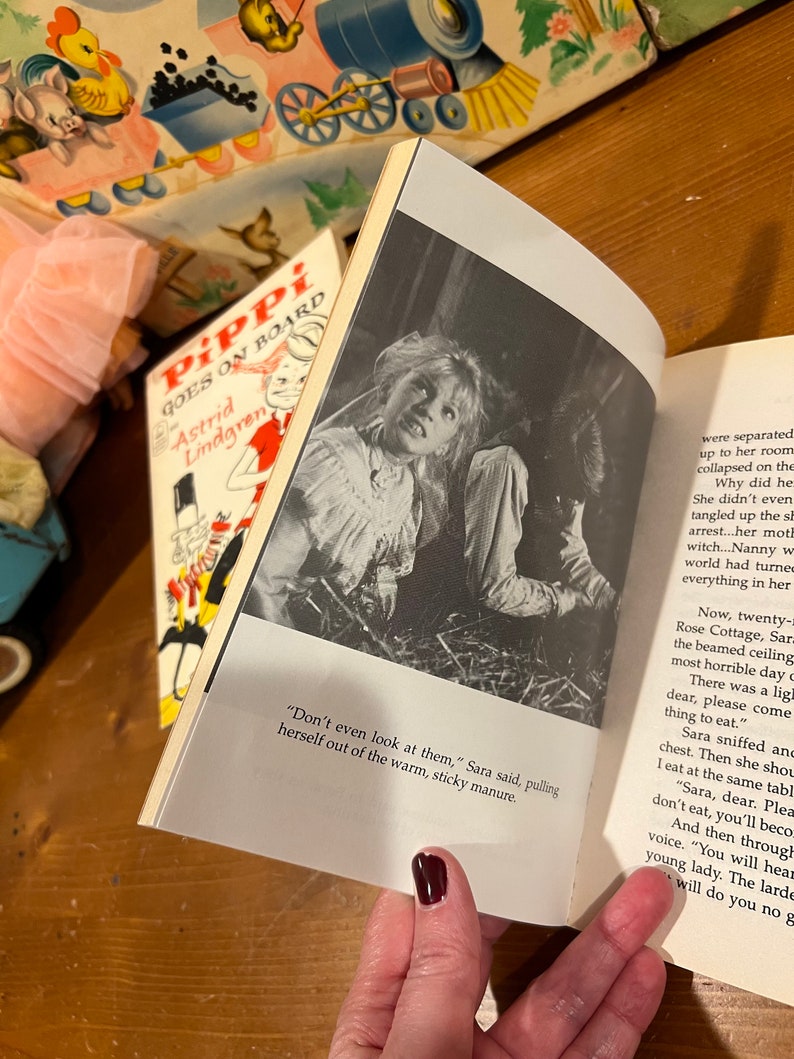 May include: A close-up of a book open to a page with a black and white photo of a young girl with a worried expression. The text on the page reads "Don't even look at them," Sara said, pulling herself out of the warm, sticky manure."