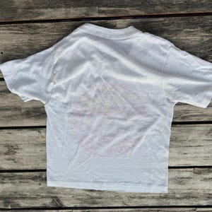May include: A plain white short-sleeved t-shirt with a crew neckline. The garment is made of a lightweight fabric and features a simple, solid white design with no visible graphics, patterns, or text on the front surface of the shirt.