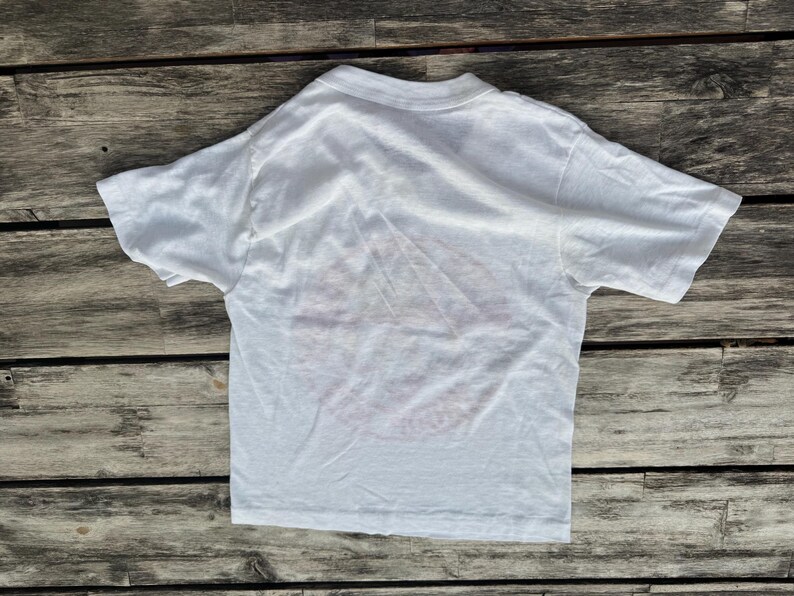 May include: A plain white short-sleeved t-shirt with a crew neckline. The garment is made of a lightweight fabric and features a simple, solid white design with no visible graphics, patterns, or text on the front surface of the shirt.