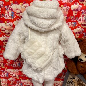 May include: A white, fluffy, hooded coat with a matching purse. The coat has a soft, textured appearance and a cozy design. The purse is attached with a thin strap. The coat is displayed on a mannequin.