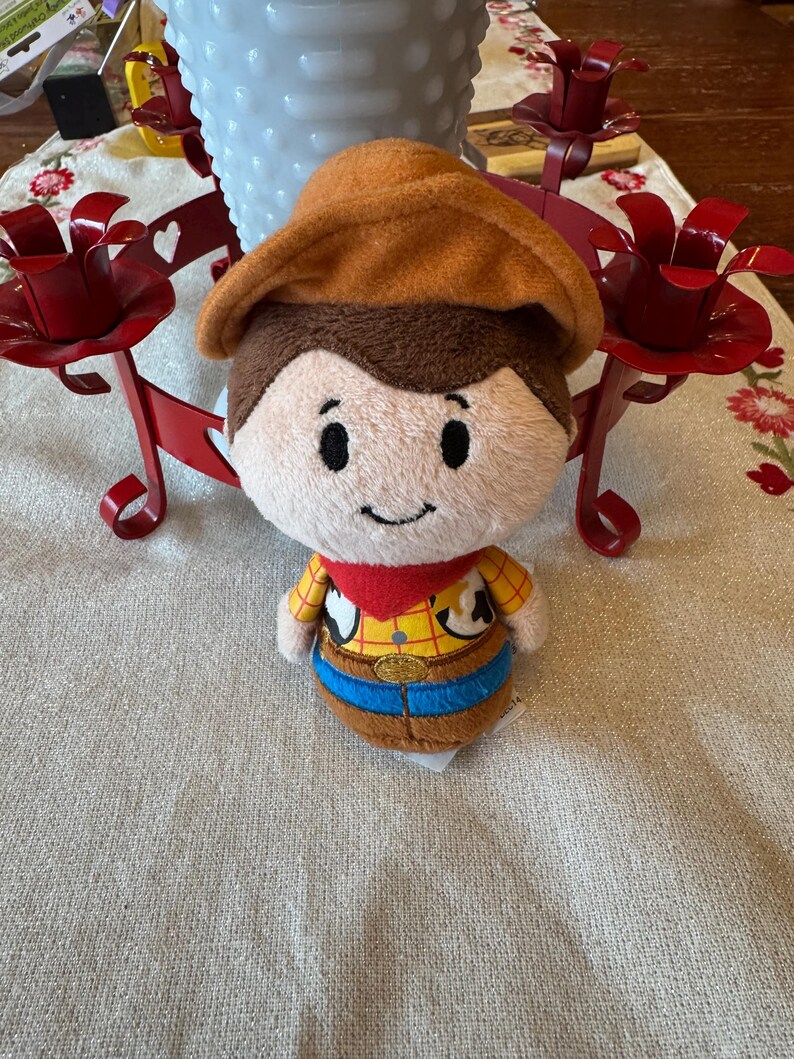 May include: A soft toy depicting a cartoon cowboy character, with a brown hat, brown hair, and a smiling face. The toy is dressed in a yellow shirt, brown vest, blue trousers, and a red neckerchief. It rests on a cream-coloured surface.