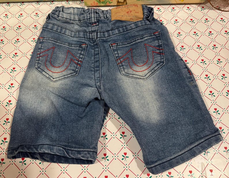 以下が含まれることがあります： バックポケットに赤いステッチが入ったブルーデニムのショートパンツ。 ショーツは色あせたウォッシュ加工で、True Religionのブランドラベルが付いています。 ショーツは伸縮性のあるウエストバンドが付いています。