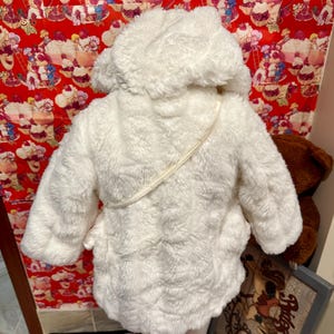 May include: A white, fluffy hooded coat with a textured surface. The coat has a cream-colored strap and a soft, plush appearance. The coat is designed for children.