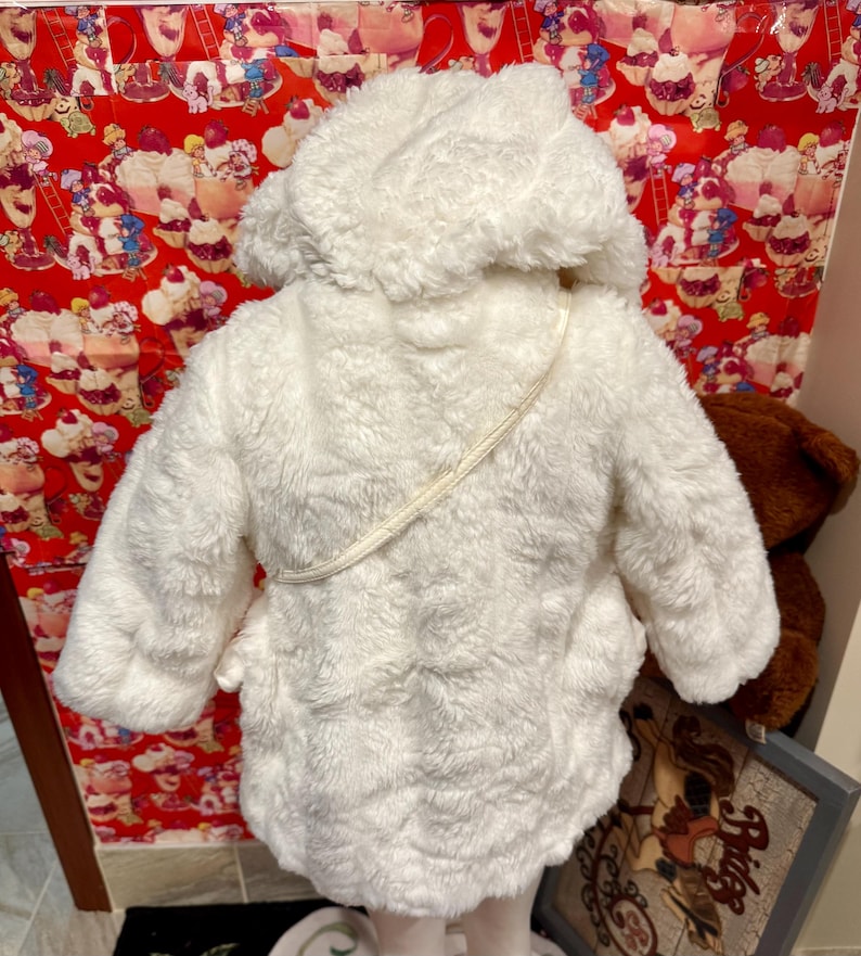 May include: A white, fluffy hooded coat with a textured surface. The coat has a cream-colored strap and a soft, plush appearance. The coat is designed for children.