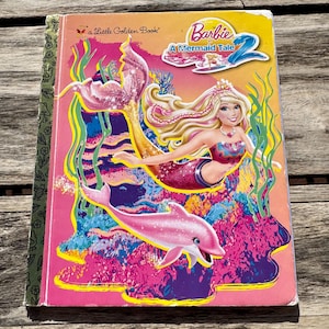Pode incluir: Um livro infantil intitulado "Barbie em Uma Hist&oacute;ria de Sereias 2". A capa apresenta uma sereia com cabelo loiro, uma cauda rosa e um golfinho rosa. O fundo &eacute; uma cena subaqu&aacute;tica colorida com tons de rosa, azul e amarelo.