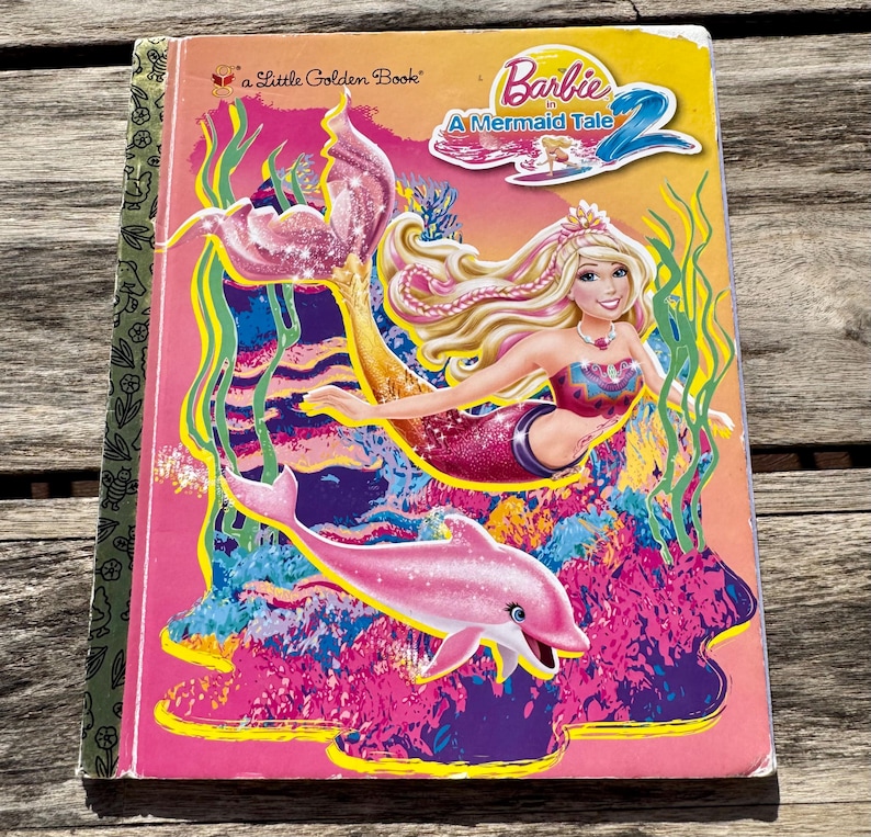 Pode incluir: Um livro infantil intitulado "Barbie em Uma Hist&oacute;ria de Sereias 2". A capa apresenta uma sereia com cabelo loiro, uma cauda rosa e um golfinho rosa. O fundo &eacute; uma cena subaqu&aacute;tica colorida com tons de rosa, azul e amarelo.