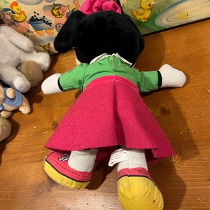 May include: A plush toy of a cartoon character wearing a green dress, red skirt, and yellow shoes. The toy has black ears and a pink bow on its head.