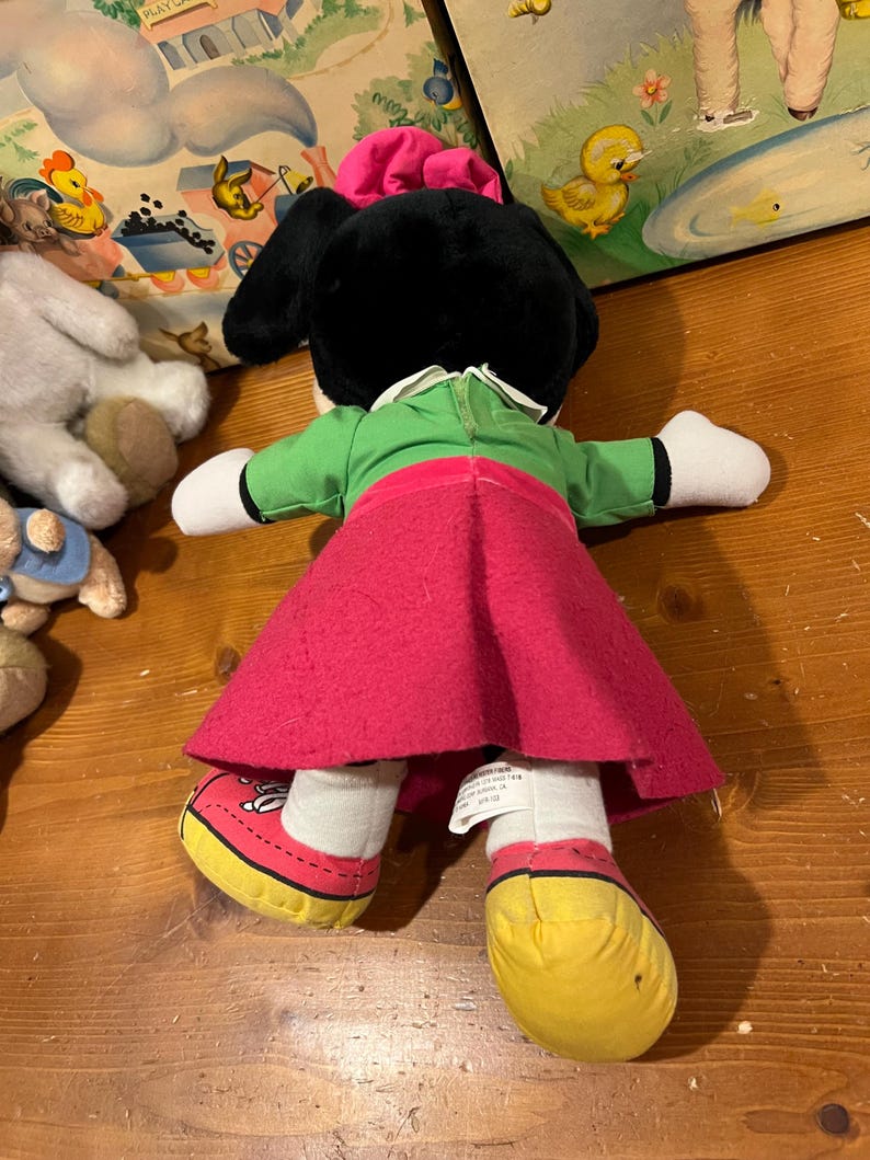 May include: A plush toy of a cartoon character wearing a green dress, red skirt, and yellow shoes. The toy has black ears and a pink bow on its head.