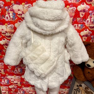 May include: A white, fluffy, hooded coat with a matching purse. The coat has a soft, textured appearance and a cozy design. The purse is attached to the coat with a thin strap. The coat is displayed on a mannequin.