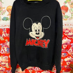 Op de afbeelding: Zwart sweatshirt met een grote Mickey Mouse afbeelding en het woord "MICKEY" in rode letters.