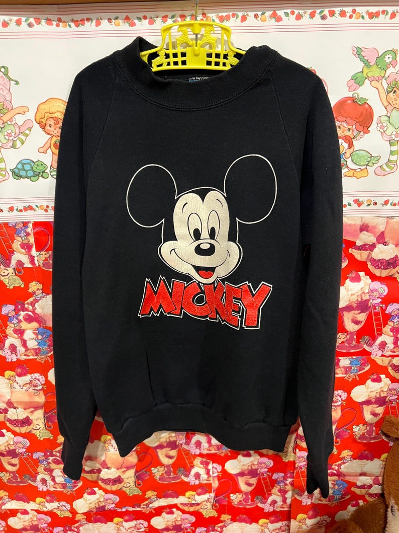 Op de afbeelding: Zwart sweatshirt met een grote Mickey Mouse afbeelding en het woord "MICKEY" in rode letters.