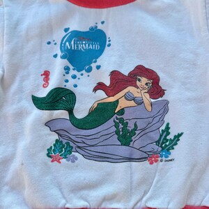 Op de afbeelding: Een wit shirt met lange mouwen en een afbeelding van Ari&euml;l de Kleine Zeemeermin die op een paarse rots rust. Het shirt heeft een groenblauwe en roze opstaande kraag en een roze zoom. De afbeelding bevat ook bubbels en het Disney-logo.