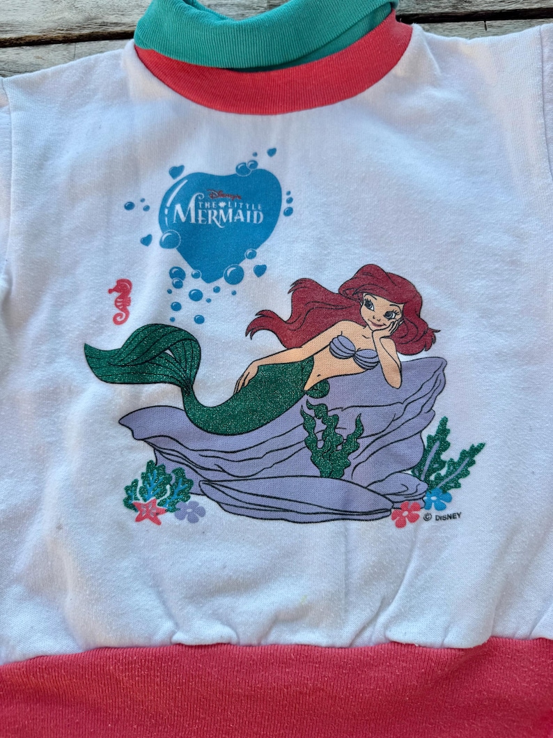 Op de afbeelding: Een wit shirt met lange mouwen en een afbeelding van Ari&euml;l de Kleine Zeemeermin die op een paarse rots rust. Het shirt heeft een groenblauwe en roze opstaande kraag en een roze zoom. De afbeelding bevat ook bubbels en het Disney-logo.