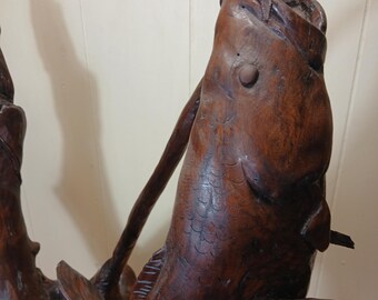 May include: A detailed, hand-carved wooden sculpture of a fish with its mouth open, appearing to be catching a wooden rod. The sculpture is a rich, dark brown color, with visible wood grain and intricate details. The fish's scales and fins are carefully rendered.