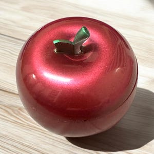 Könnte beinhalten: Ein glänzender, roter Zierapfel mit Metallic-Finish. Der Apfel hat einen grünen Stiel und ein grünes Blatt. Das Objekt hat eine solide, runde Form und liegt auf einer hellen Holzoberfläche.
