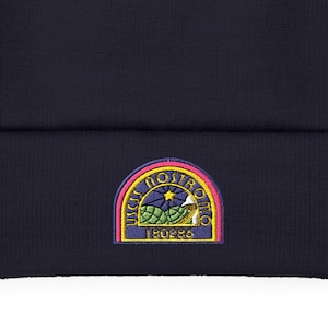 May include: A navy blue beanie with a colorful embroidered patch. The patch features a rainbow arch with text and a floral design. The beanie is a classic, comfortable style, suitable for various occasions.